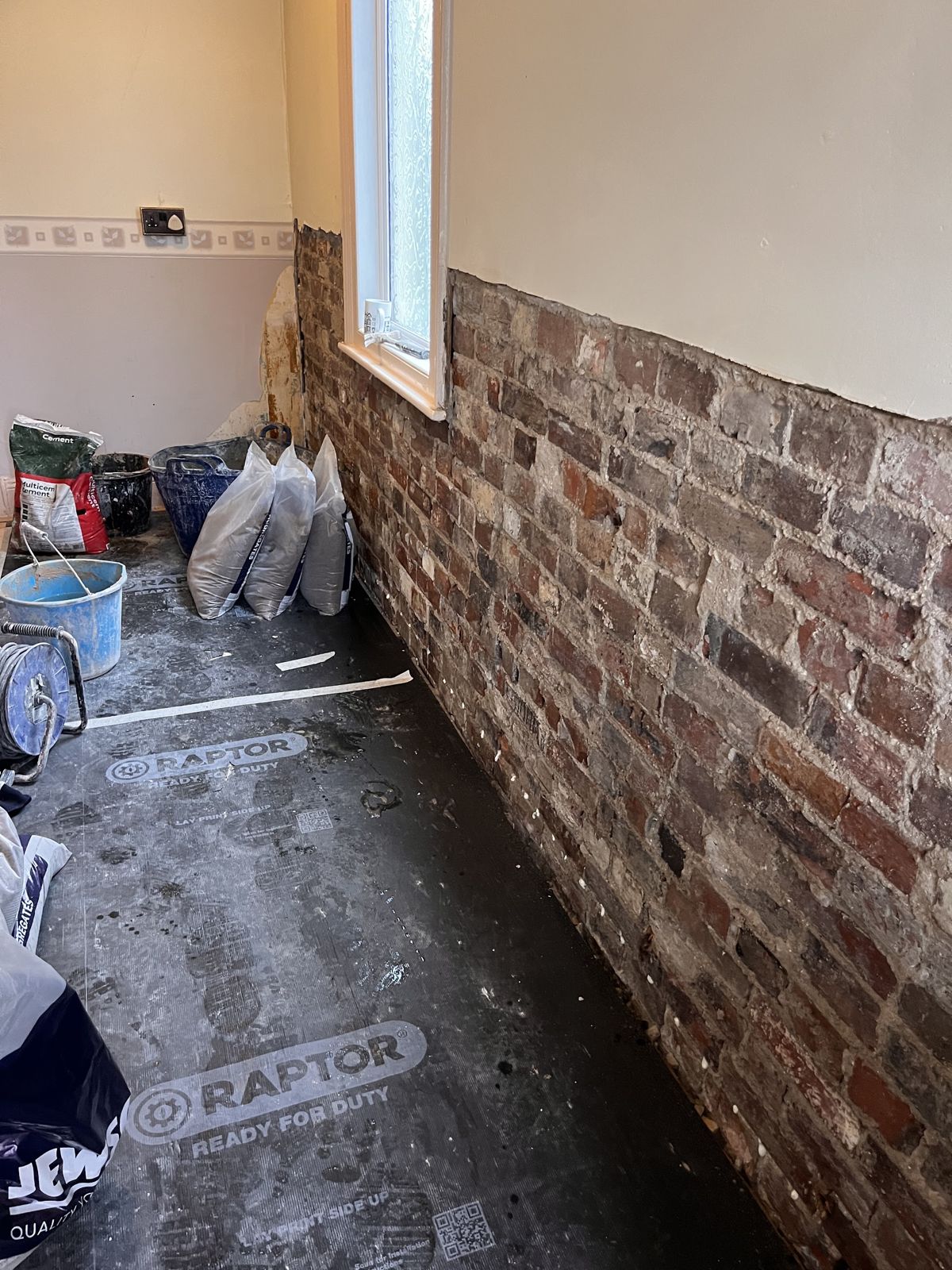 Plaster stripped to bare brick along the lower part of a wall under a window, with bags of materials on a protected floor